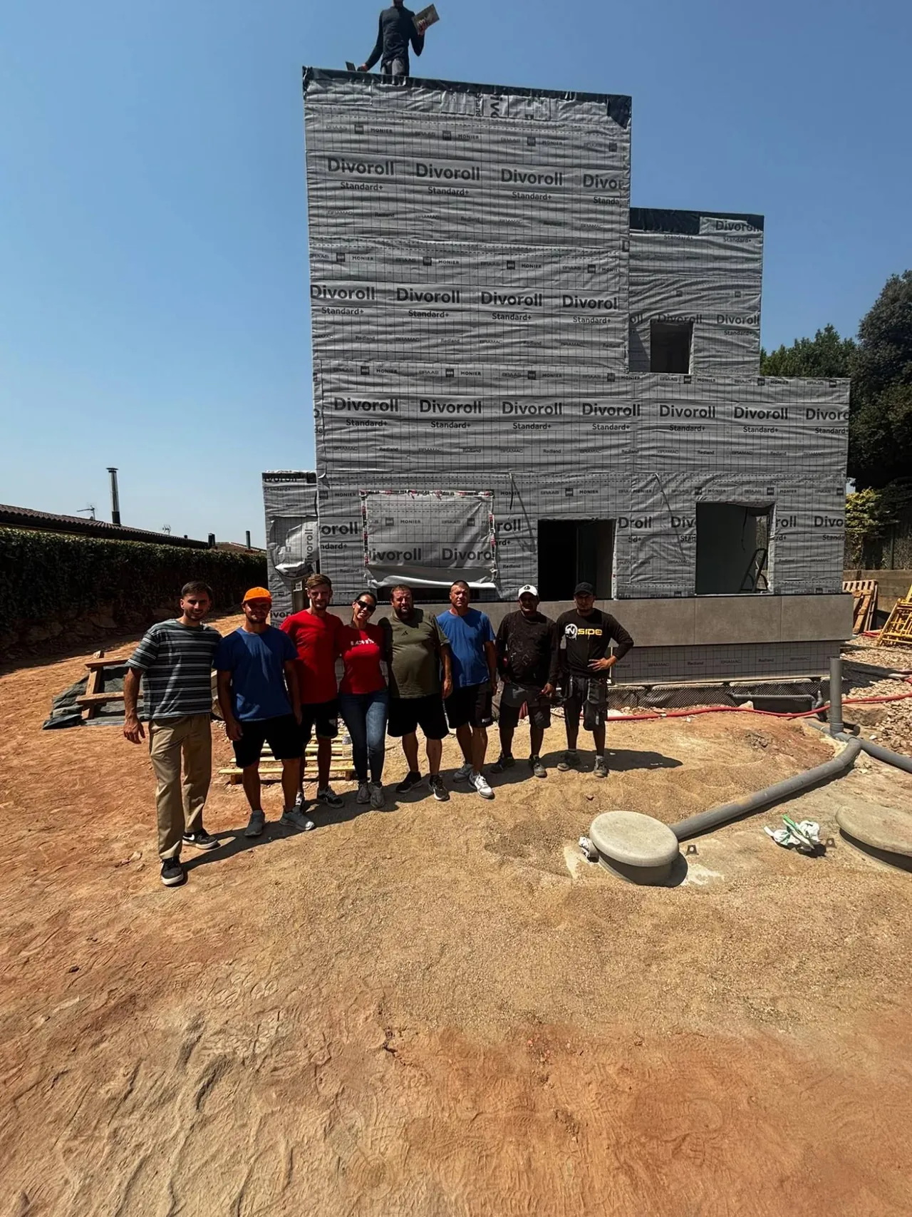 Equip d'obra davant d'un habitatge en construcció, amb la façana coberta per la làmina de protecció.